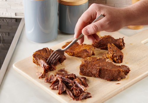 A person shredding cooked beef on a cutting board using a fork.