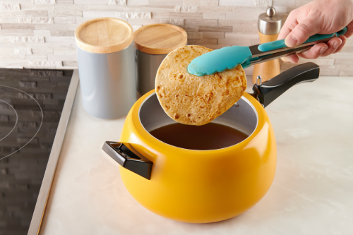 A hand is seen dipping a tortilla into a steaming pot of seasoned broth over a 10-inch nonstick skillet on medium heat.